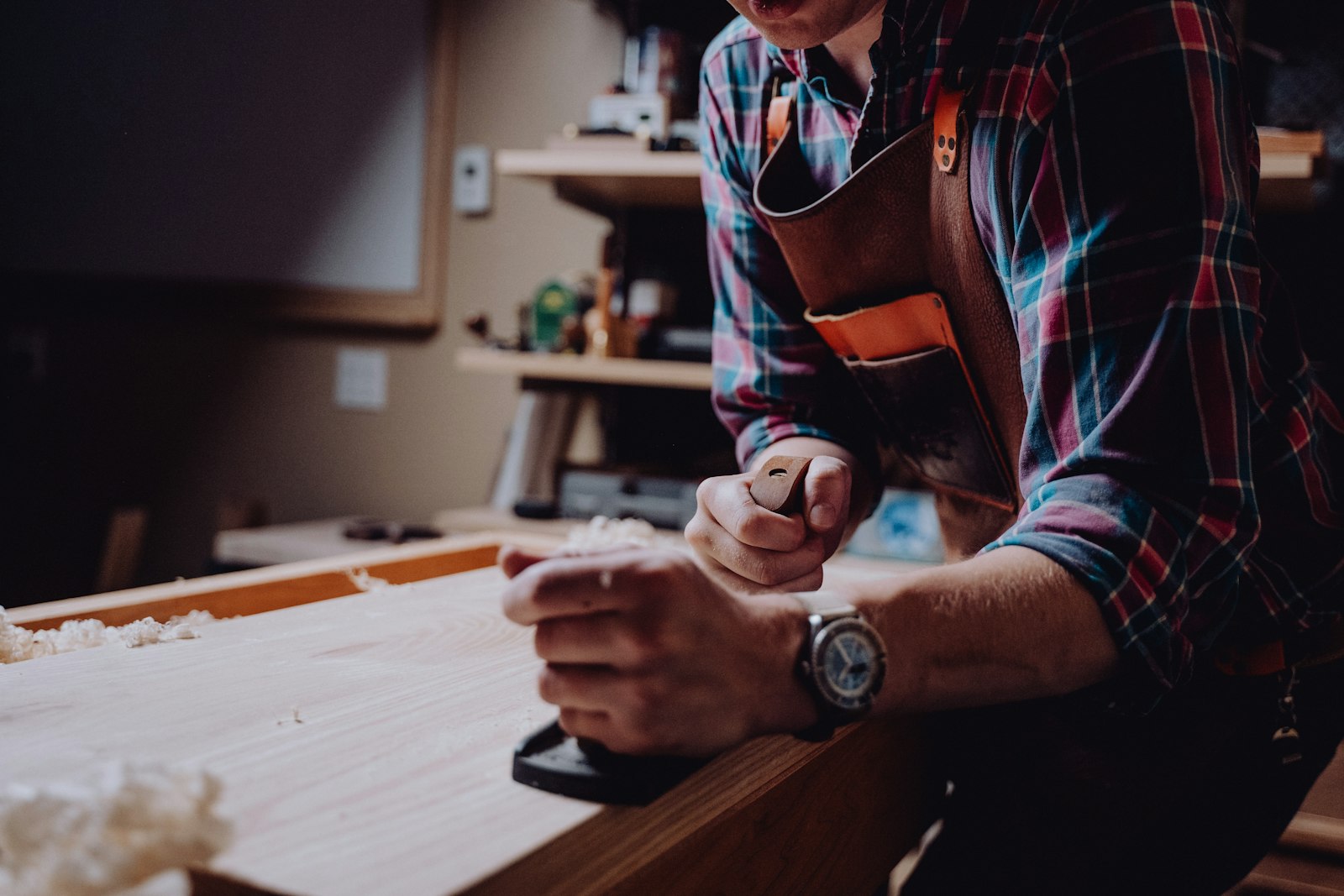 Un artisan rabote une planche de bois à la main, tablier de cuir noué à la taille.