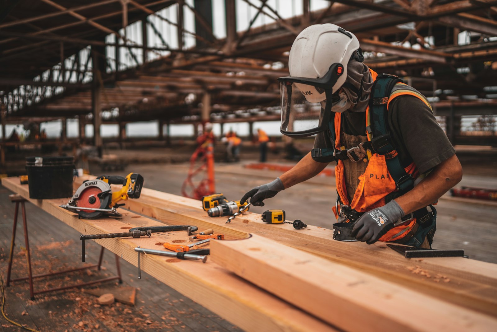 Un ouvrier équipé d'un casque et d'une visière assemble une poutre de bois avec des outils électroportatifs.