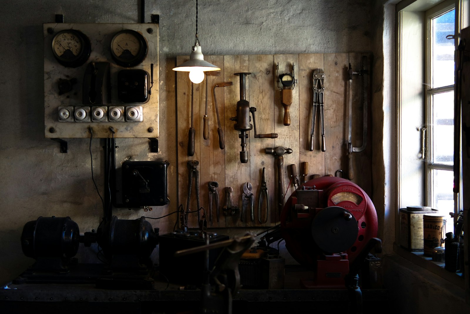 Panneau mural d'outils de mécanicien anciens, pinces et manivelle, sous une lampe suspendue.